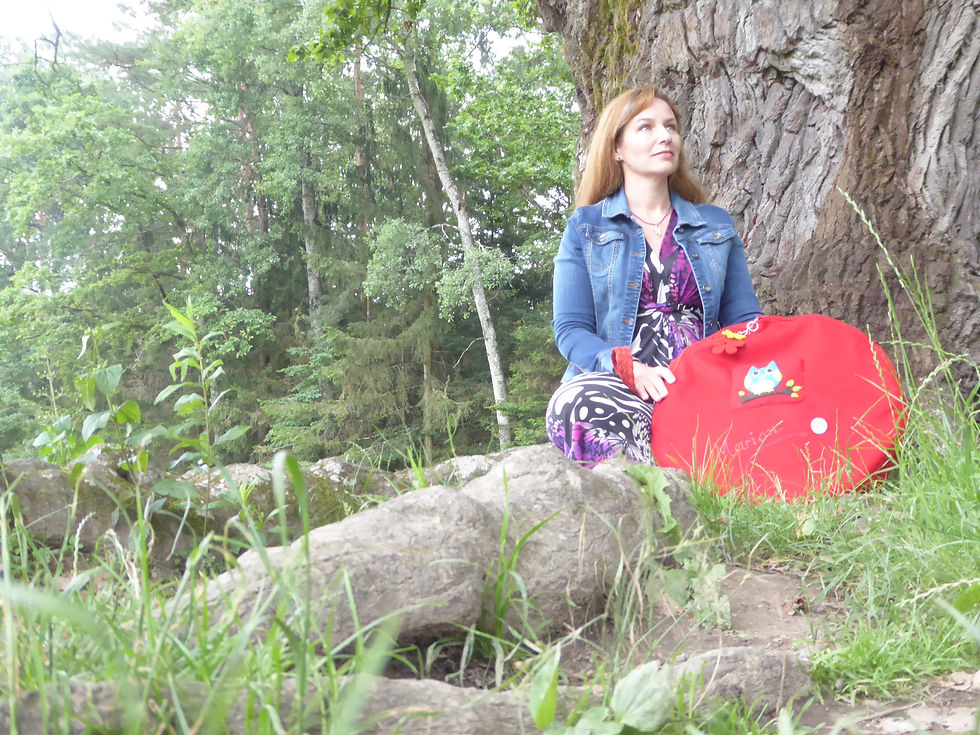 Frau sitzt nachdenklich an einem großen Baumstamm im Grünen, hält eine rote Trommel und ist umgeben von Steinen, Gras und Waldlandschaft.