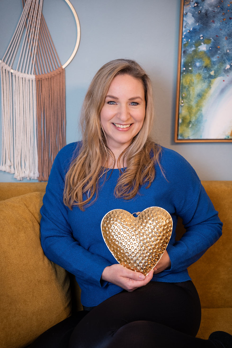 Marion Siener sitzt lächelnd auf einem Sofa und hält ein goldenes Herz in den Händen – Symbol für gelebte Herzkraft und Herzensarbeit