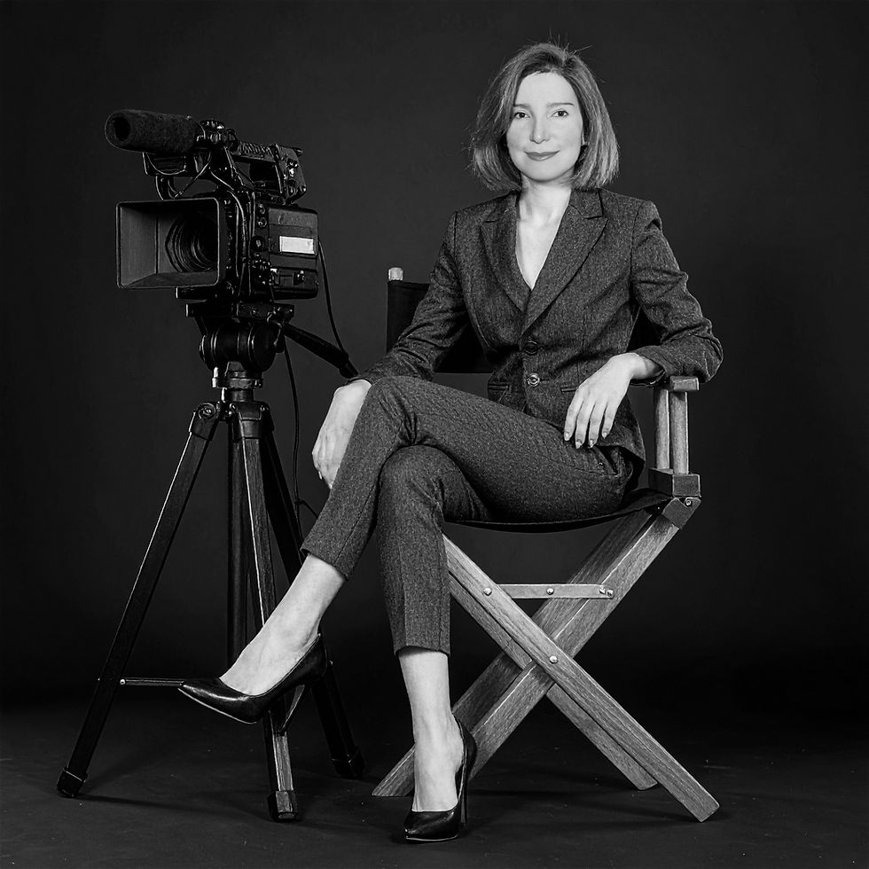 A woman in a suit sits confidently on a director's chair beside a professional video camera against a dark background. Black-and-white image.