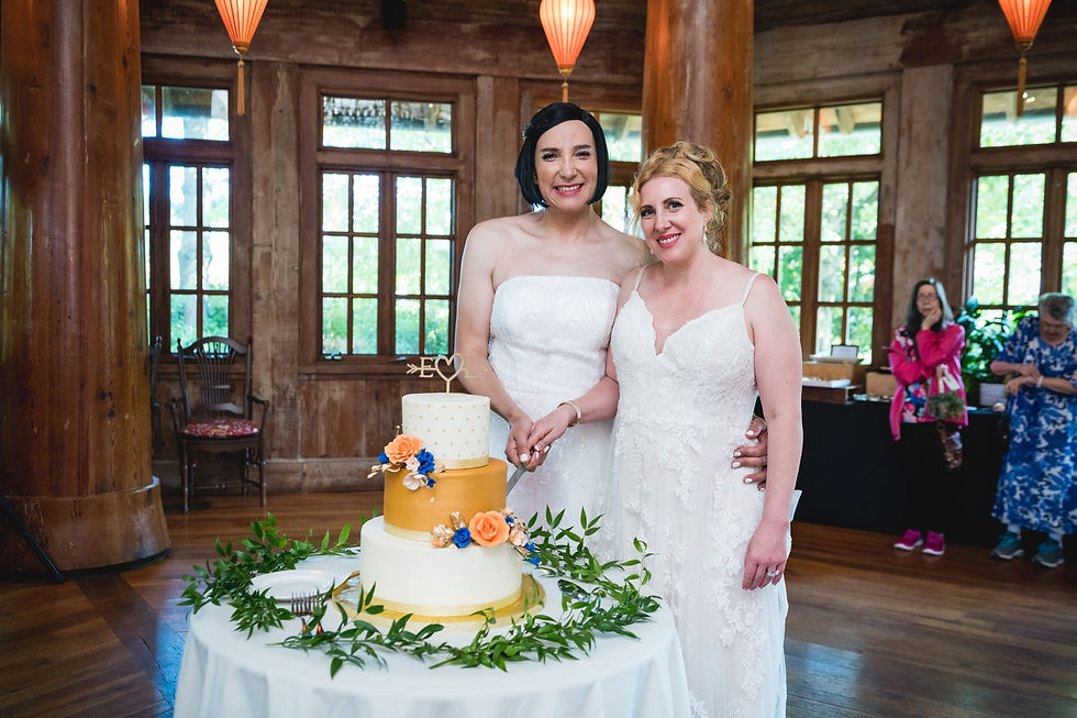 Two brides in white dresses smile, posing with a decorated wedding cake in a wood-paneled room. Guests and greenery are in the background.
