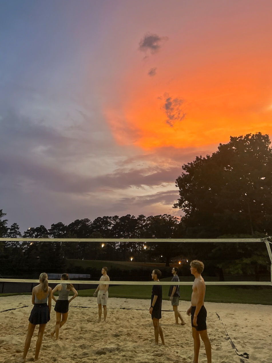 People play beach volleyball at sunset under a vibrant orange sky, surrounded by trees. The mood is relaxed and focused.