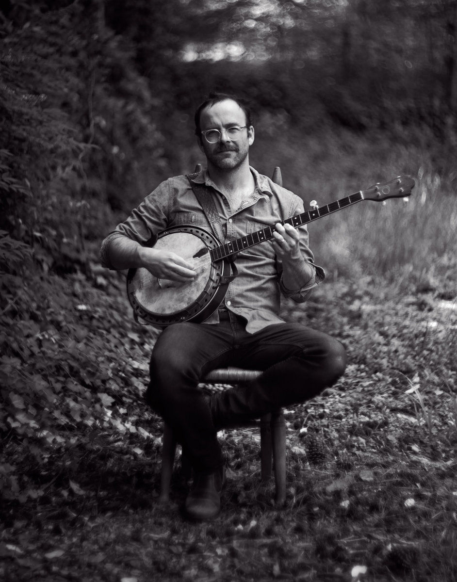Joseph Decosimo sits outdoors on a chair playing a banjo, wearing glasses and a casual shirt. The background is a blurred forest, creating a serene mood.