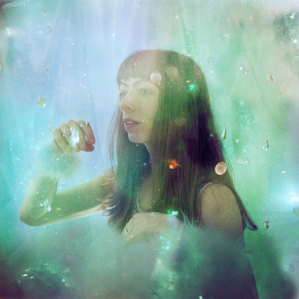 The cover for Elsa Hewitt's Chaos Emeralds album, showing Hewitt in ethereal green-blue setting, surrounded by glistening particles, looks contemplative with hand raised. Dreamy and serene mood.