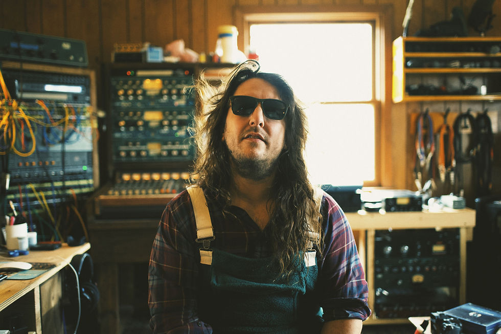 An unshaven Leroi Conroy with long hair, dungarees and sunglasses sits in a recording studio. Background has analogue audio equipment and cables. Warm, relaxed atmosphere.