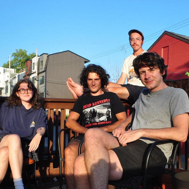 The band Vireo sitting outside on a deck, smiling. One wears a "Dead Kennedys" shirt. Bright, sunny day with colorful buildings in the background.