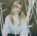 Brighton based folk artist Rachel Adderley aka REA, in a white top sits amid dry grass, gazing calmly at the camera in a soft, muted outdoor portrait.