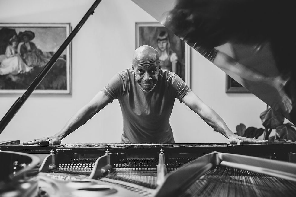 Laraaji smiling over grand piano, leaning in with hands on the frame. Black and white photo with paintings in the background.