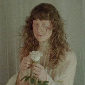 The cover for Lucy Kruger & The Lost Boys', Anchor single, showing Lucy Kruger with long hair holding a white rose, eyes closed. Soft lighting and muted colors create a serene, introspective mood.