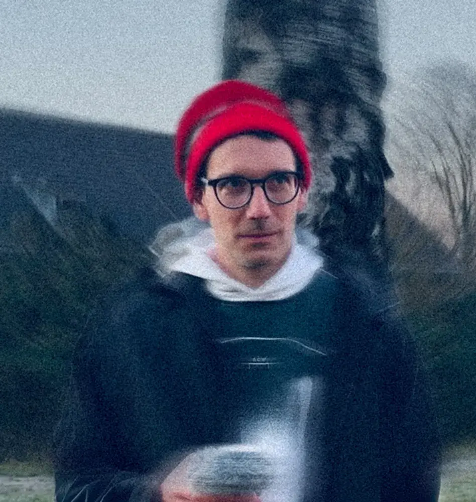 Cologne based producer and composer, Boris Rogowski in red beanie and glasses holds a fuzzy object, standing outdoors with blurred background of trees and a building, creating a dreamy effect.