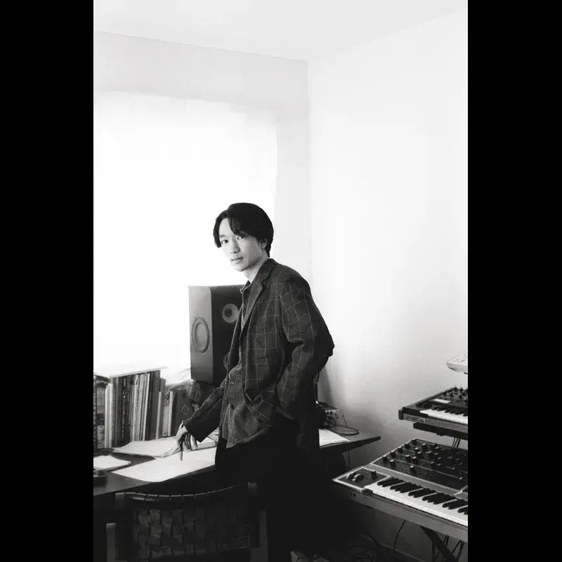 Black-and-white photo of Japanese producer and musician Yuma Koda in a music studio, standing by a desk with records and synth keyboards, looking calmly at camera