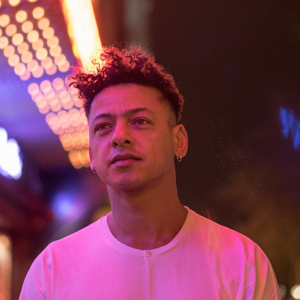 Portable Aka Bodycode with curly hair in a white shirt stands under neon lights, gazing thoughtfully. The atmosphere is moody and colorful.