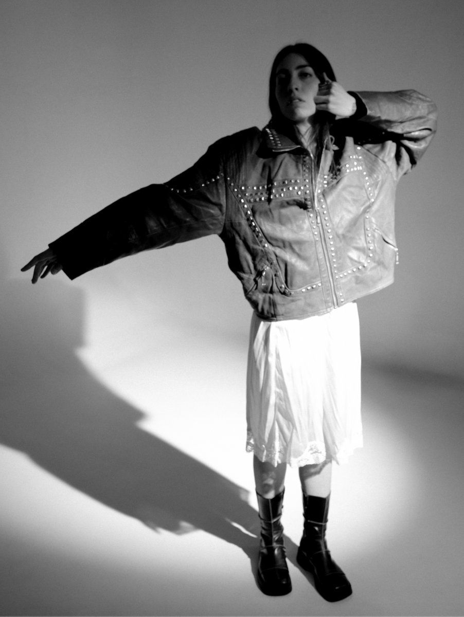 Black-and-white studio portrait of Mali Obomsawin posing in an oversized studded jacket, white skirt, and black boots, with a long shadow.