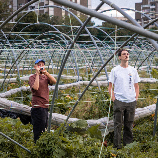 Electronic music due, Orange and Mountains, in a greenhouse framework; one drinks from a mug, the other looks around. Urban buildings in the background; relaxed atmosphere.