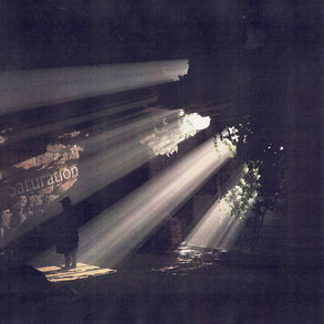 The cover for Arbee's Saturation album, showing sunbeams filtering through a dark, dilapidated room with plants. A silhouette stands amidst debris. Text "Saturation" is partially visible.