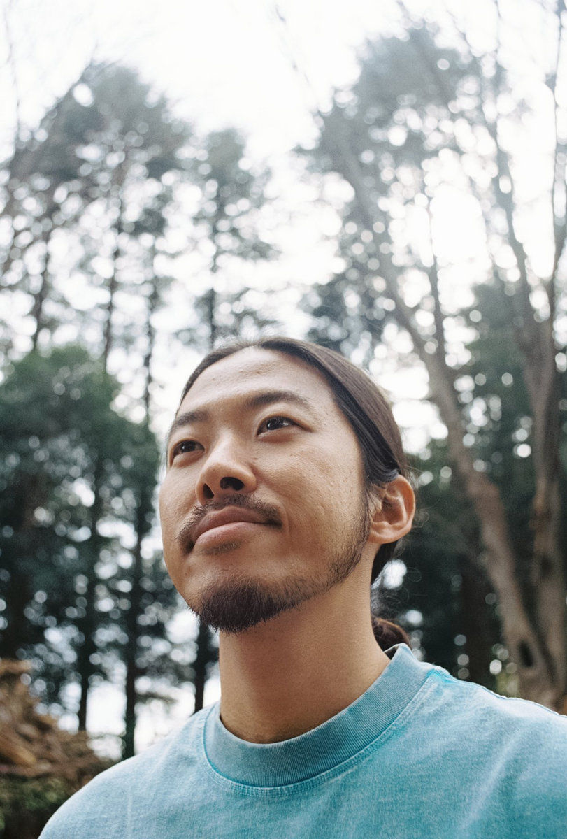 Close-up of Yuji Namiki aka nubo, in a blue shirt looking upward in a misty forest, calm and reflective.