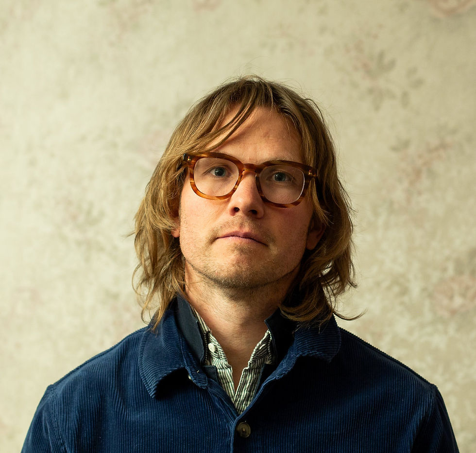 Jens Kuross with glasses and long hair, wearing a blue jacket and striped shirt, stands against a floral beige wall, looking calmly forward.
