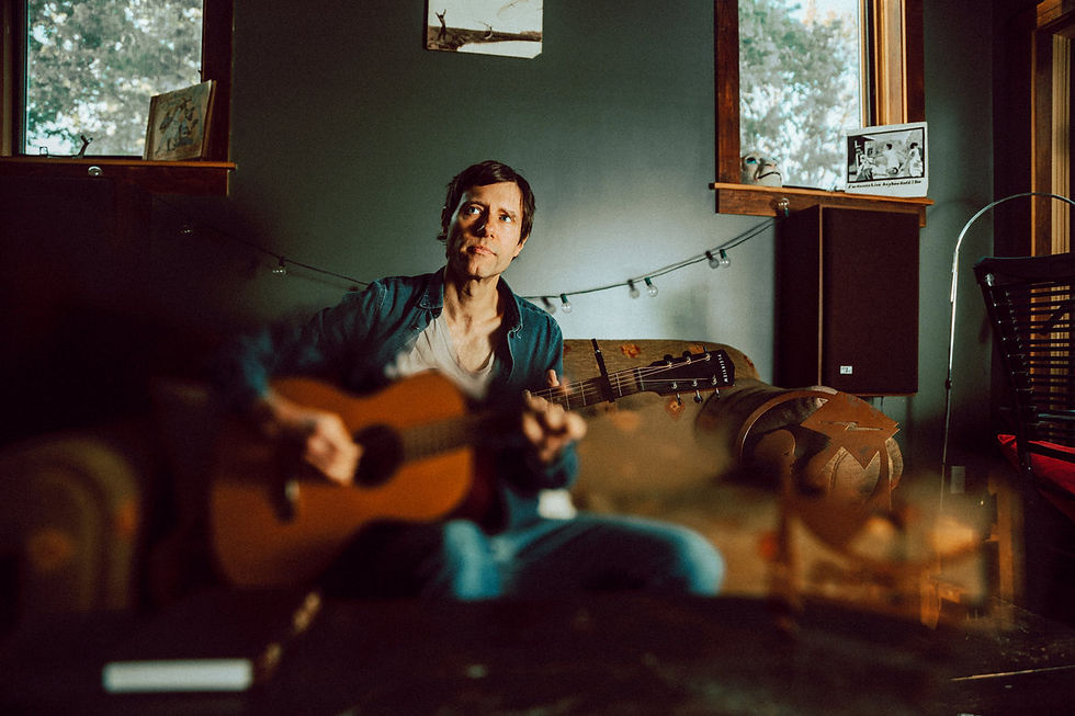 Nathan Salsburg playing guitar in a cozy room with dim lighting. String lights and framed photos on walls create a relaxed, reflective mood.