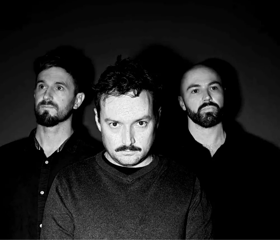 Dale Hardman, Todd James and Tyson Mahoney, aka Geomancer, with serious expressions stand closely together in a dark setting. The image is in black and white, with strong shadows.