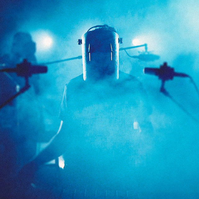 Composer and sound designer Tim Harrison, aka Recur, in a helmet plays music on a smoky, blue-lit stage, surrounded by microphones. The mood is mysterious and atmospheric.