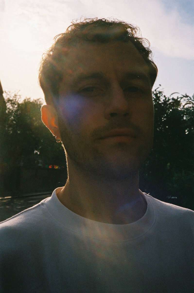 Backlit photo of Ogul Can Genc aka SONLIFE in a white shirt outdoors, sun flare behind him, with trees in the background and a serious expression.