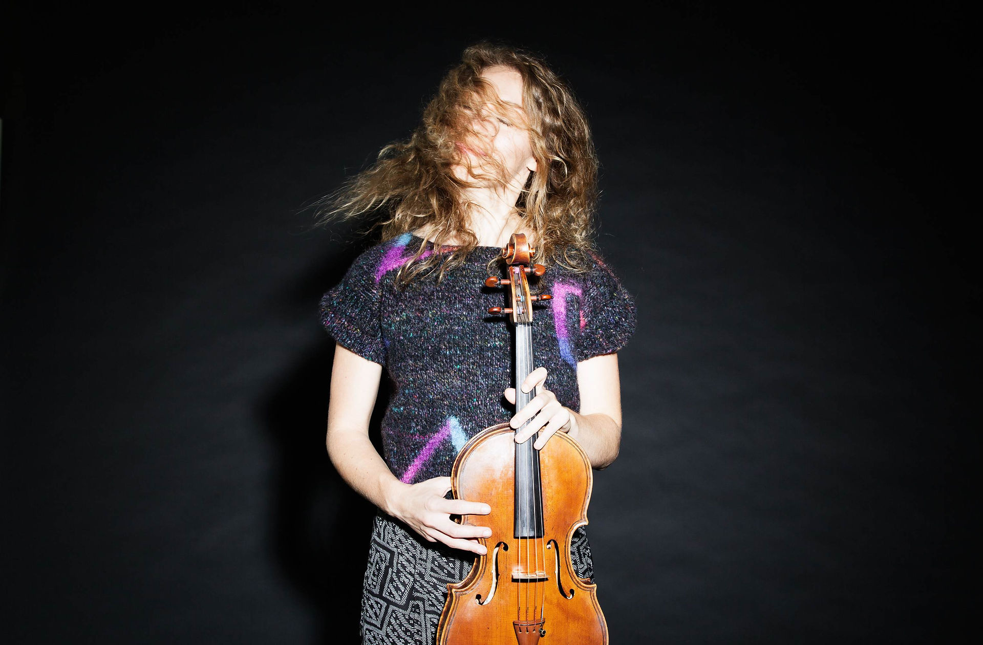 A slightly blurry press shot of Marta Sofia Honer holding her violin upright with her hair blowing over her face partially obscuring it. The background is black.