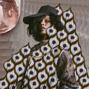 This is the cover for Perera Elsewhere’s Just Wanna Live Some album, showing Elsewhere wearing colourful, peacock feather patterned attire and a black cap stands against a textured background, exuding a bold and confident mood.