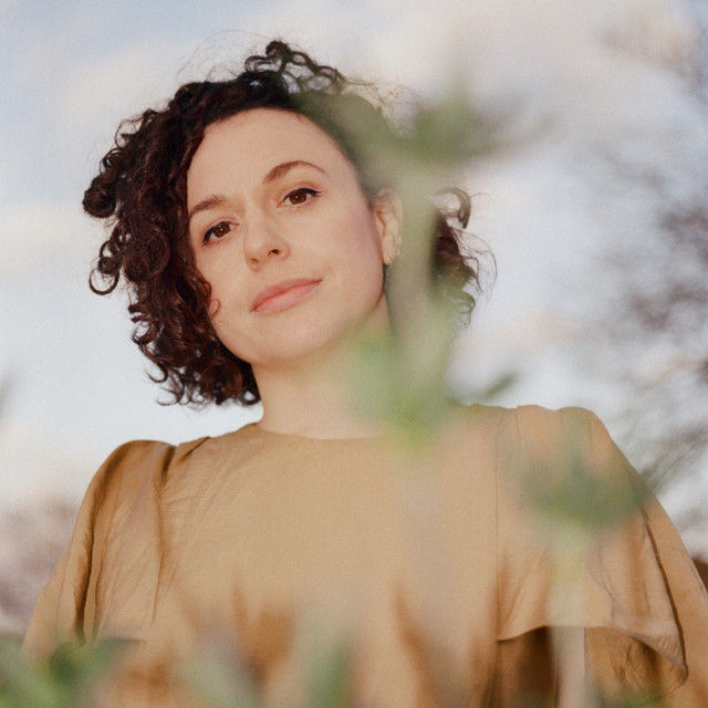 Portrait of Nomieye aka Rosie Turton, with curly dark hair in a tan dress, seen through blurred foliage against a soft sky, calm expression.