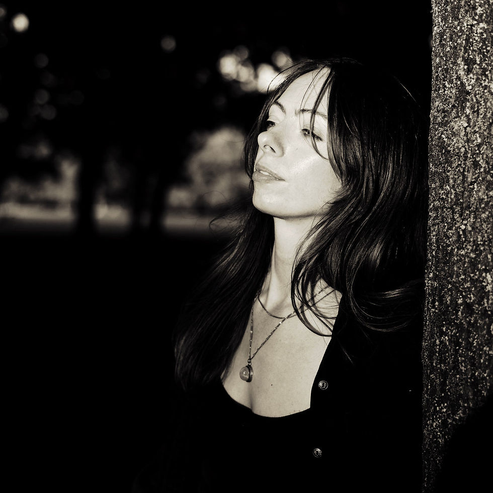 Elsa Hewittwith long hair leans against a tree, gazing thoughtfully. Sepia tone. Blurred trees in the background suggest a park setting.