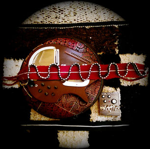 The cover for Evan Golde aka Goldstar's Zig Zag album, showing a circular leather foot rest with paisley design, red and black strap with an exotic stringed instrumement on it and a strap with beaded detail. "Solstice Electrics" text on packaging nearby. Black and white background.