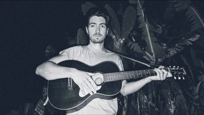 Montreal-based, multi-instrumentalist Clay Hazey holding a guitar in a tropical background at night. He wears a light shirt and appears calm. Dark foliage adds a moody atmosphere.
