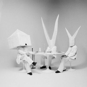 The cover for Crayon's Home Safe album, showing three figures in white suits sit at a round table with tall paper hats. The setting is minimalistic and monochromatic, creating a surreal mood.