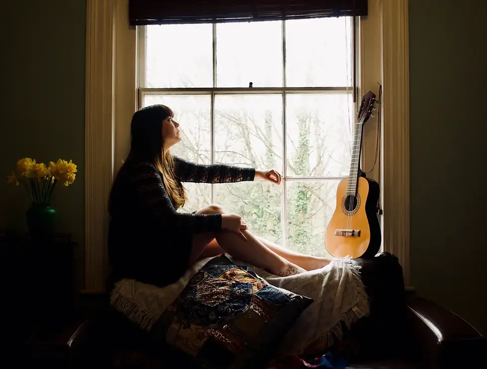 Folk singer Lucy Kitchen sits on a windowsill, gazing outside. A guitar rests nearby. Daffodils in a vase and patterned cushions add color to the cozy room.