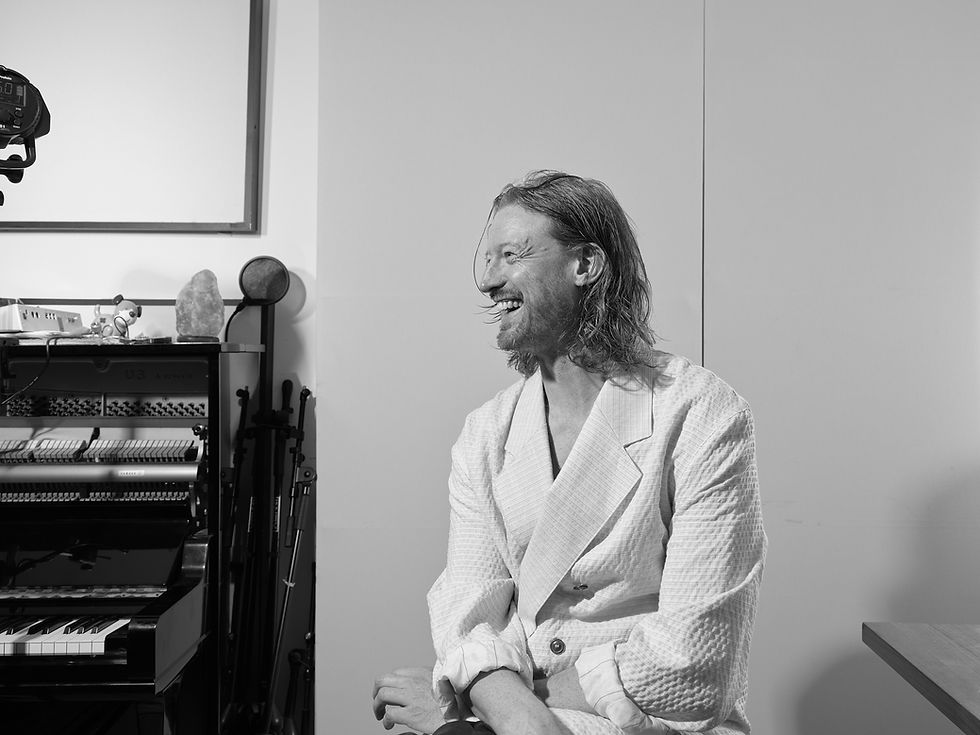 Black-and-white portrait of Dutch ambient artist Andert Tysma in a textured jacket, seated beside a piano and camera gear in a studio