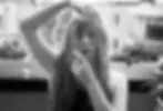 Black-and-white portrait of Kristina Cox aka Selkie, outdoors, one arm over her head and a finger near her lips, with potted plants behind her.