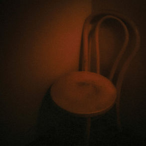 The cover for Lawrence English & Stephen Vitiello's Trinity album, showing a wooden chair with a curved back, dimly lit in a shadowy corner, creating a moody and mysterious atmosphere with warm brown tones.