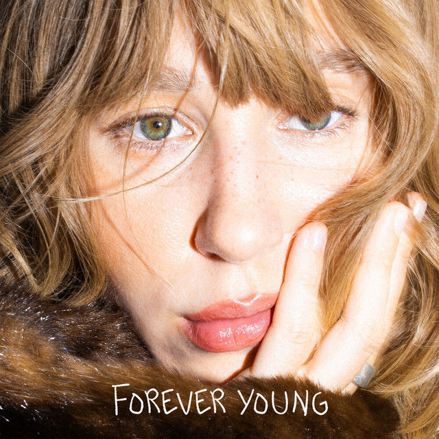 Close-up of a Marie Noreger with light brown hair and green eyes, hand on cheek, wearing fur. Text "Forever Young" - the title of her latest single in white, conveying a reflective mood.