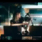 Leva Vaiti aka Piksel, playing violin into a microphone in a dim studio, focused and calm, with laptops and audio gear in the foreground.