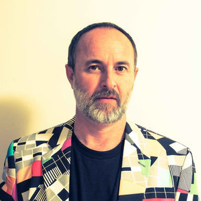Lee Nicholson AKA Test Card, with a beard in a colorful, geometric-patterned jacket stands against a light background. Neutral expression, minimal shadows.