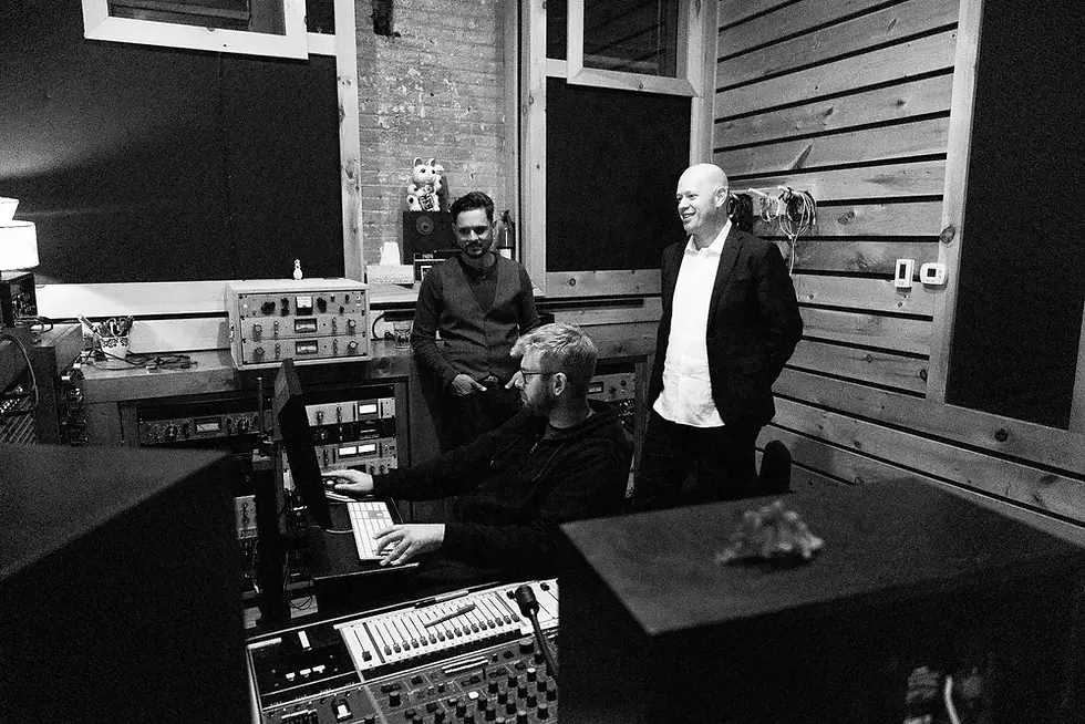 Ben Cockerham of The Royal Pacific Orchestra with two others in a recording studio, one at a computer and two standing nearby, surrounded by audio equipment in black and white.