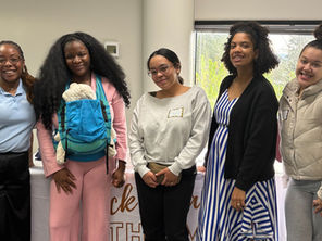 Cinco mujeres negras están juntas y sonríen a la cámara. Una de ellas lleva a su bebé en un portabebés azul.