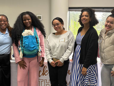 Cinco mujeres negras están juntas y sonríen a la cámara. Una de ellas lleva a su bebé en un portabebés azul.