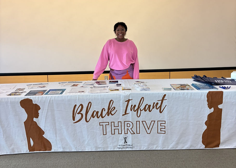 A woman with black hair wearing a pink sweatshirt smiles behind a table that says "Black Infant Thrive"