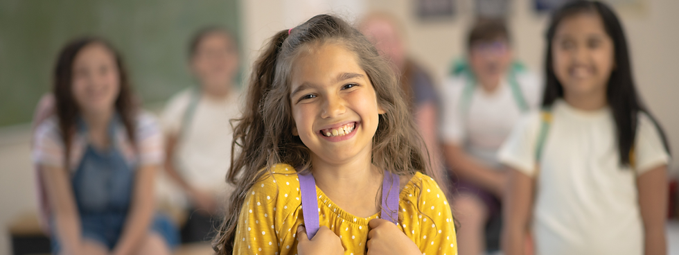A young girl at school, holding the straps of her backpack and smiling widely.