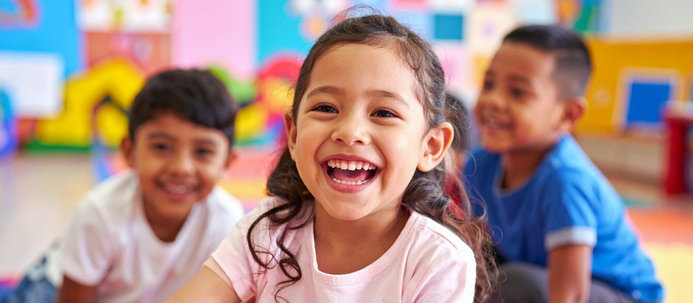 A young girl in school smiles widely. Two young boys are smiling behind her.