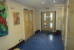 A hallway with blue flooring, cream walls, double doors, artwork, and cleaning equipment.