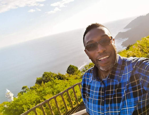 Black smiling man, Michael Johnson, in glasses and a blue checked shirt, taking a selfie with a scenic coastline in the backg