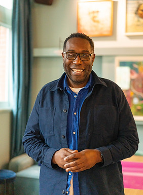 Black smiling man, Michael Johnson, smiling indoors and wearing glasses with colourful artwork in the background