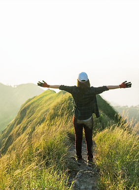 Person standing on a grassy hilltop with arms outstretched, facing the sunrise