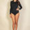 Thumbnail: Woman standing and posing in a long-sleeve black bodysuit against a plain light-colored background.