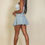 Thumbnail: A woman with long dark hair stands sideways, wearing a white tank top and a light blue pleated mini skirt.